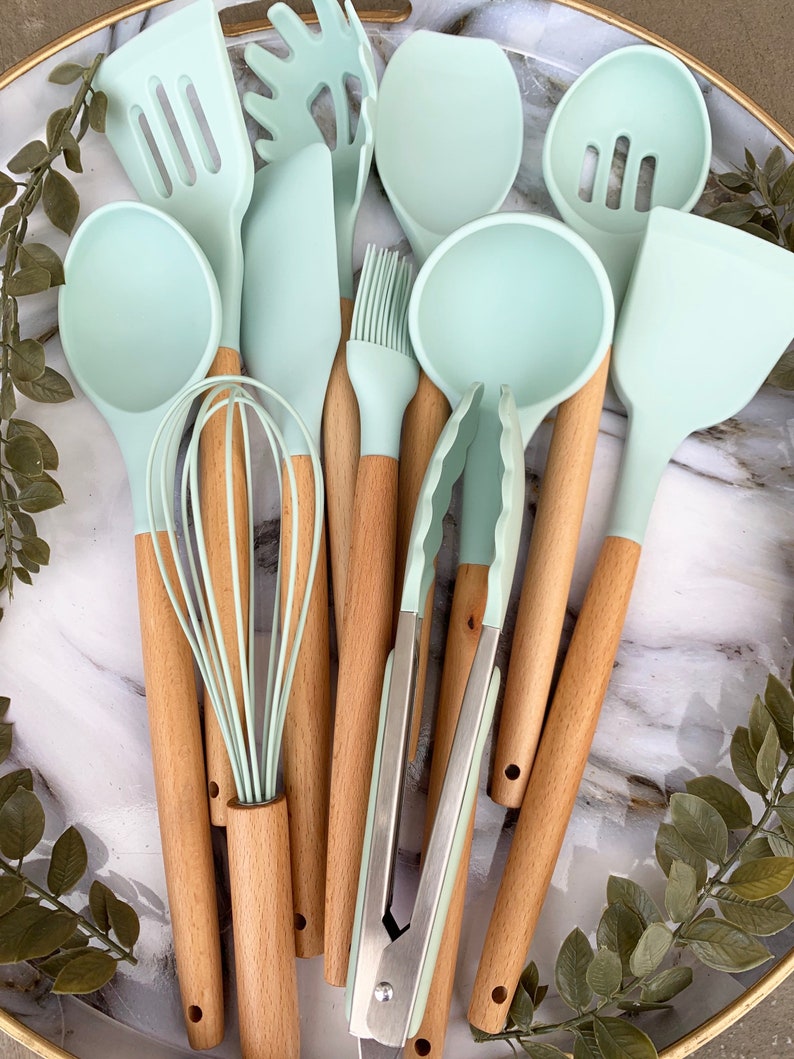 May include: A set of ten kitchen utensils with light blue silicone heads and wooden handles. The utensils include a spatula, a spoon, a whisk, a ladle, a slotted spoon, a basting brush, a set of tongs, and a spatula with a scalloped edge.
