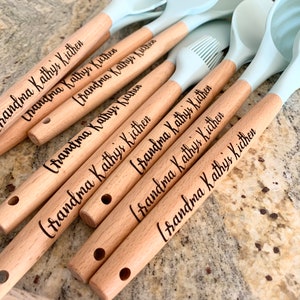 May include: A set of six wooden handled kitchen utensils with blue silicone heads. The handles are engraved with the text "Grandma Kathy's Kitchen".