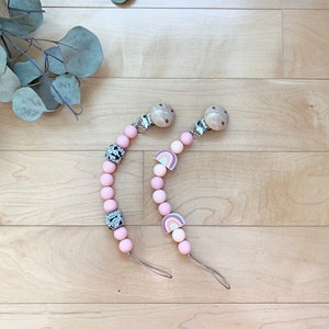 Peut inclure: Deux anneaux de dentition pour bébé en silicone rose et blanc avec des clips en bois. Un anneau de dentition a un motif arc-en-ciel et l'autre a un motif géométrique noir et blanc.