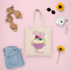 May include: A cream-colored tote bag featuring a cupcake design with a pink frosting, berries, and sprinkles. The bag is surrounded by a pair of brown sandals, sunglasses, a white camera, a pair of blue jeans, and sunflowers on a pink background.