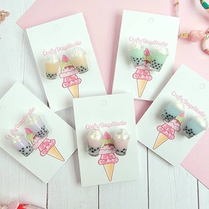 May include: Four pairs of bubble tea earrings on white card stock with the text "Crafty Days Studio". The earrings are in four different colors: purple, pink, green, and blue. Each pair of earrings is a miniature version of a bubble tea cup with a white lid and a straw.