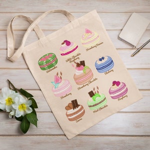 May include: A beige tote bag featuring colorful illustrations of various desserts, including watermelon, strawberry, and blueberry macarons. The bag has a simple design with two handles and is placed on a wooden surface.