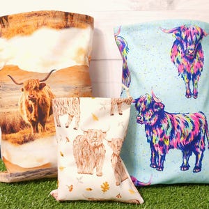Puede incluir: Tres bolsas de tela con dise&ntilde;os de vacas de las Highlands. Una bolsa presenta una vaca realista en un paisaje, otra tiene un fondo azul claro con vacas coloridas, y la tercera tiene un fondo crema con ilustraciones de vacas y hojas de oto&ntilde;o.