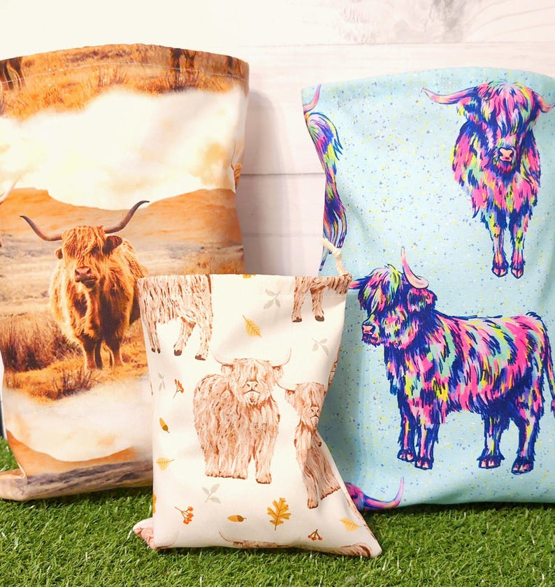 Puede incluir: Tres bolsas de tela con dise&ntilde;os de vacas de las Highlands. Una bolsa presenta una vaca realista en un paisaje, otra tiene un fondo azul claro con vacas coloridas, y la tercera tiene un fondo crema con ilustraciones de vacas y hojas de oto&ntilde;o.
