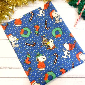 May include: Blue fabric with a repeating Christmas-themed pattern. The design features Snoopy characters in Santa hats and sweaters, candy canes, wreaths, and Christmas lights. The background is covered in small white and yellow dots.