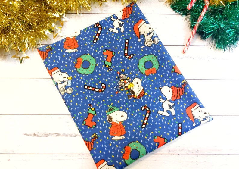 May include: Blue fabric with a repeating Christmas-themed pattern. The design features Snoopy characters in Santa hats and sweaters, candy canes, wreaths, and Christmas lights. The background is covered in small white and yellow dots.