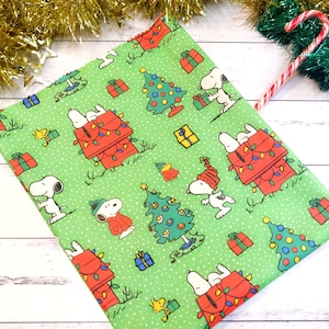 May include: Green fabric featuring Snoopy and Woodstock in a Christmas theme. Snoopy is shown in various poses with a red doghouse decorated with Christmas lights, Christmas trees, and wrapped gifts.
