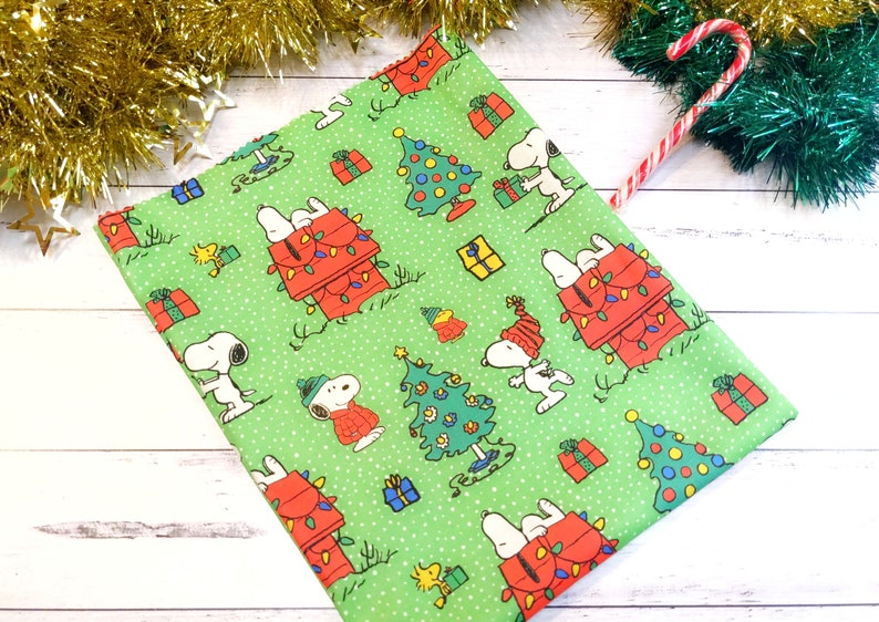May include: Green fabric featuring Snoopy and Woodstock in a Christmas theme. Snoopy is shown in various poses with a red doghouse decorated with Christmas lights, Christmas trees, and wrapped gifts.