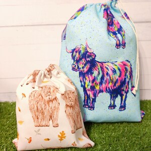 two reusable fabric cow bags. One Autumn themed, the other has multicoloured cows in a bohemian style fabric on a blue background. both bags are sitting on green grass with a white wooden background