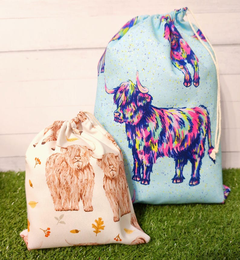 two reusable fabric cow bags. One Autumn themed, the other has multicoloured cows in a bohemian style fabric on a blue background. both bags are sitting on green grass with a white wooden background