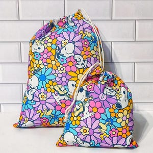 May include: Two drawstring bags with a colorful floral and cartoon character pattern. The bags feature a repeating design of flowers in shades of pink, purple, yellow, and blue, along with cartoon characters. The bags have white drawstrings.