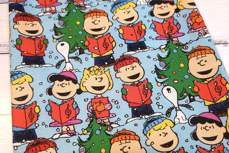 May include: Fabric featuring a repeating pattern of cartoon characters singing carols, with Christmas trees and Snoopy on a light blue background. The characters are wearing hats and holding red songbooks. The design is festive and colorful.