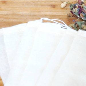 May include: Six white muslin tea bags with a drawstring closure. The bags are folded and stacked on a wooden surface.