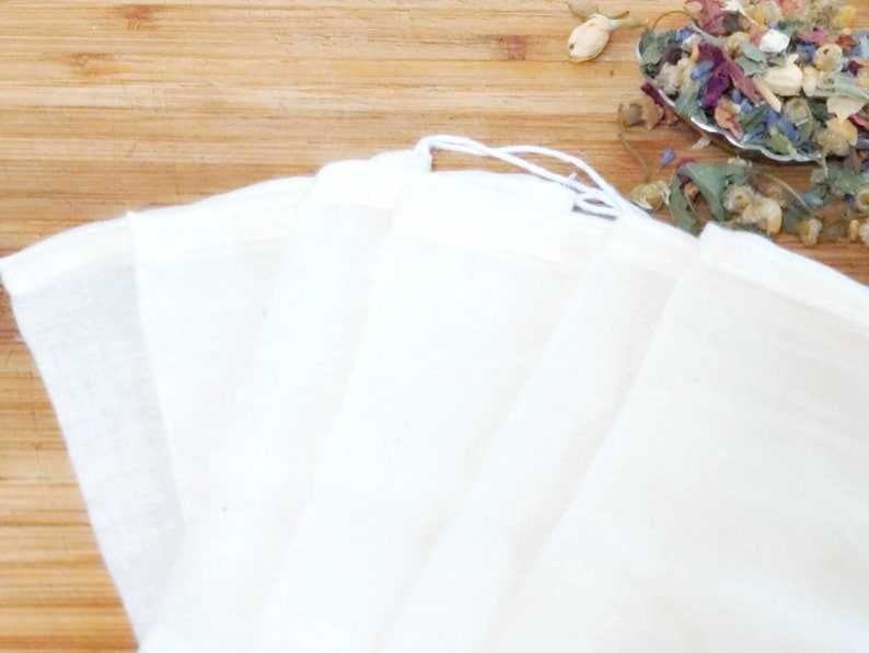 May include: Six white muslin tea bags with a drawstring closure. The bags are folded and stacked on a wooden surface.