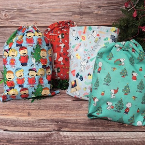 May include: Four drawstring bags with Christmas-themed patterns. One bag is blue with cartoon characters and Christmas trees. Another is red with Snoopy. A third is light blue with Snoopy and other designs. The last is teal with Snoopy and Christmas trees.