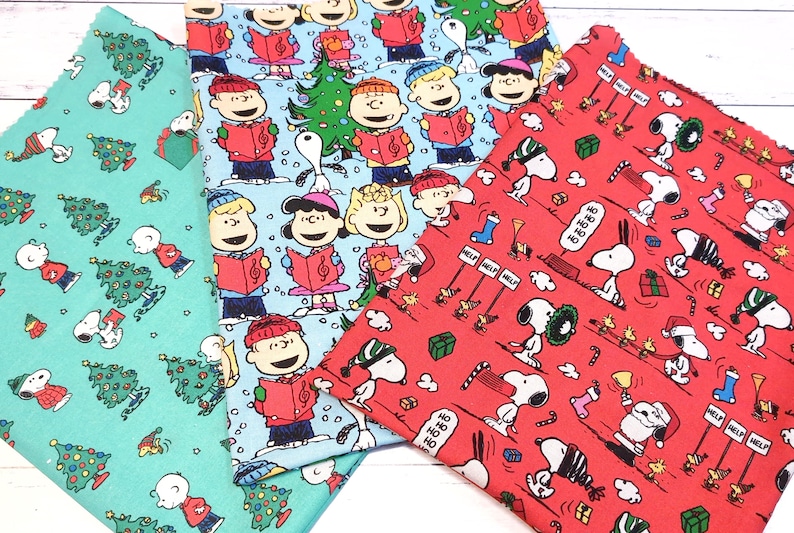 May include: Three fabric swatches with Christmas-themed patterns. One is teal with Snoopy, Christmas trees, and Charlie Brown. Another is light blue with the Peanuts gang singing carols. The third is red with Snoopy and holiday elements.