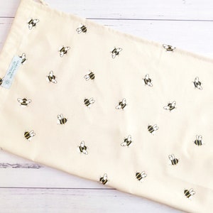 K&ouml;nnte beinhalten: Eine beige wiederverwendbare Einkaufstasche mit einem Muster aus schwarzen und gelben Bienen. Die Tasche hat einen Kordelverschluss und ein kleines Etikett mit der Aufschrift "The Good Life" und einem Sonnenmotiv.