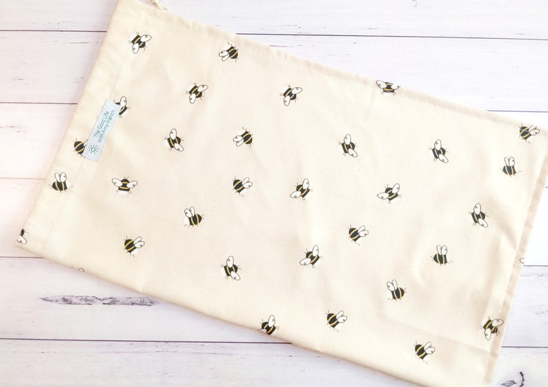 K&ouml;nnte beinhalten: Eine beige wiederverwendbare Einkaufstasche mit einem Muster aus schwarzen und gelben Bienen. Die Tasche hat einen Kordelverschluss und ein kleines Etikett mit der Aufschrift "The Good Life" und einem Sonnenmotiv.