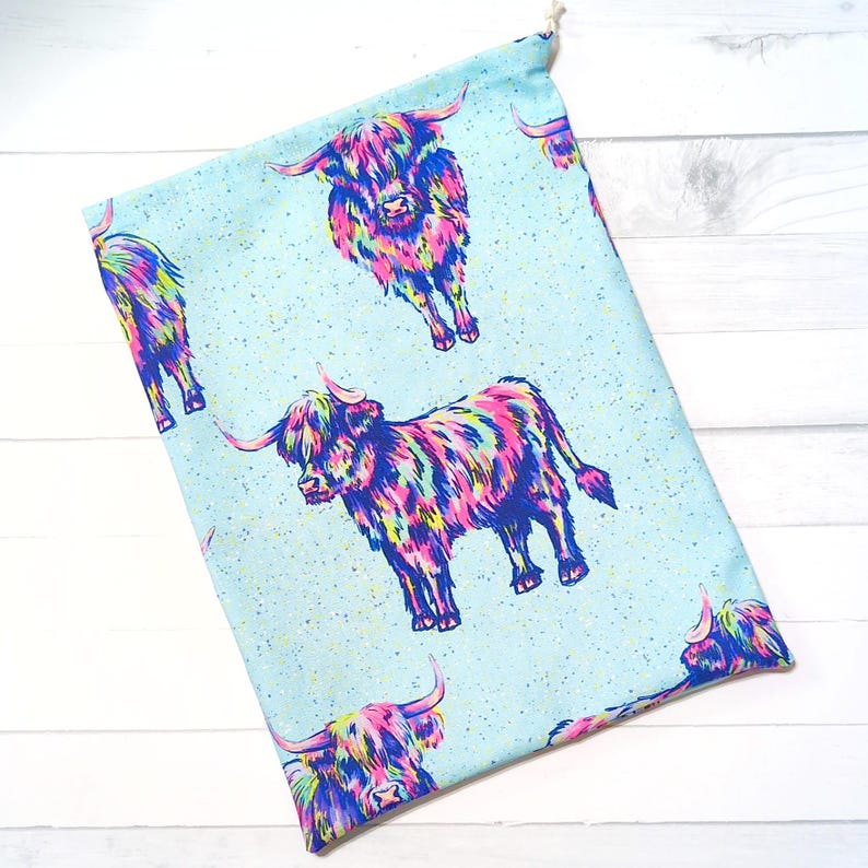 Puede incluir: Una bolsa de tela azul claro con un estampado repetido de vacas de las Tierras Altas coloridas. Las vacas est&aacute;n representadas en tonos de rosa, morado, amarillo y azul, sobre un fondo moteado. La bolsa tiene un cierre de cord&oacute;n.
