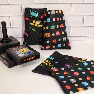 May include: Four black fabric pouches with different retro video game designs. The pouches feature Pac-Man, Space Invaders, and a cherry and ghost design. The pouches are on a white tiled surface.