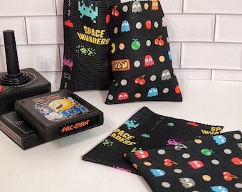 May include: Four black fabric pouches with different retro video game designs. The pouches feature Pac-Man, Space Invaders, and a cherry and ghost design. The pouches are on a white tiled surface.