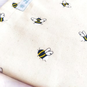 K&ouml;nnte beinhalten: Eine wei&szlig;e Stofftasche mit einem sich wiederholenden Muster aus schwarzen und gelben Bienen. Die Tasche hat ein kleines Etikett mit dem Text "Treat Yourself" darauf.