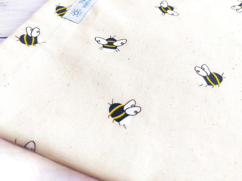 K&ouml;nnte beinhalten: Eine wei&szlig;e Stofftasche mit einem sich wiederholenden Muster aus schwarzen und gelben Bienen. Die Tasche hat ein kleines Etikett mit dem Text "Treat Yourself" darauf.