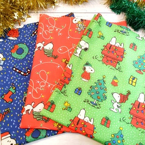 May include: Three Christmas-themed fabric pieces featuring Snoopy and Woodstock characters. The fabrics are blue, red, and green, with patterns of Snoopy, Christmas trees, and holiday lights. Festive and colorful.