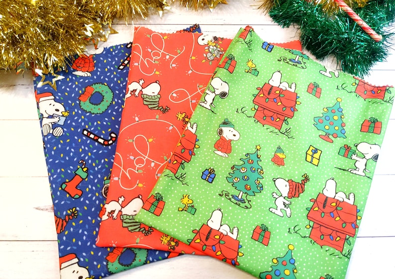 May include: Three Christmas-themed fabric pieces featuring Snoopy and Woodstock characters. The fabrics are blue, red, and green, with patterns of Snoopy, Christmas trees, and holiday lights. Festive and colorful.
