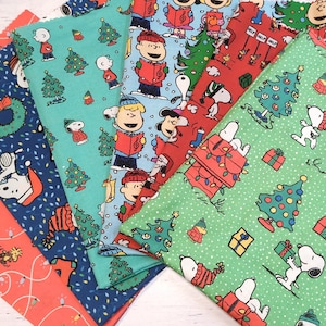 May include: A collection of Christmas-themed fabric pieces featuring Snoopy, Charlie Brown, and other Peanuts characters. The fabrics display festive patterns with Christmas trees, gifts, and holiday lights in red, blue, and green.