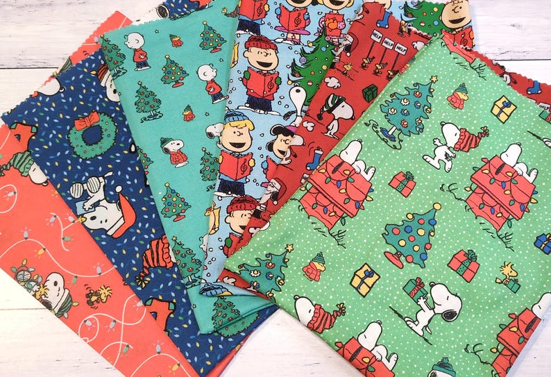 May include: A collection of Christmas-themed fabric pieces featuring Snoopy, Charlie Brown, and other Peanuts characters. The fabrics display festive patterns with Christmas trees, gifts, and holiday lights in red, blue, and green.