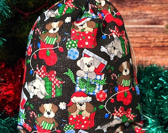 May include: A black drawstring gift bag with a festive Christmas dog pattern. The fabric features various dog illustrations, wrapped gifts, ornaments, and stockings in red, green, and white. The bag is tied with a natural-colored drawstring.