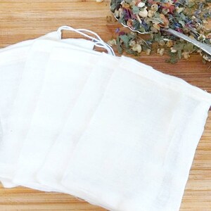 May include: Four white muslin tea bags with drawstring closures, lying flat on a wooden surface.