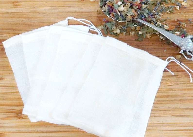 May include: Four white muslin tea bags with drawstring closures, lying flat on a wooden surface.