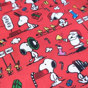 May include: Red fabric with a repeating pattern of Snoopy and Woodstock characters in Christmas scenes. Snoopy is wearing a Santa hat and scarf. The fabric also features candy canes, gifts, and the words "Ho Ho Ho" and "Help".