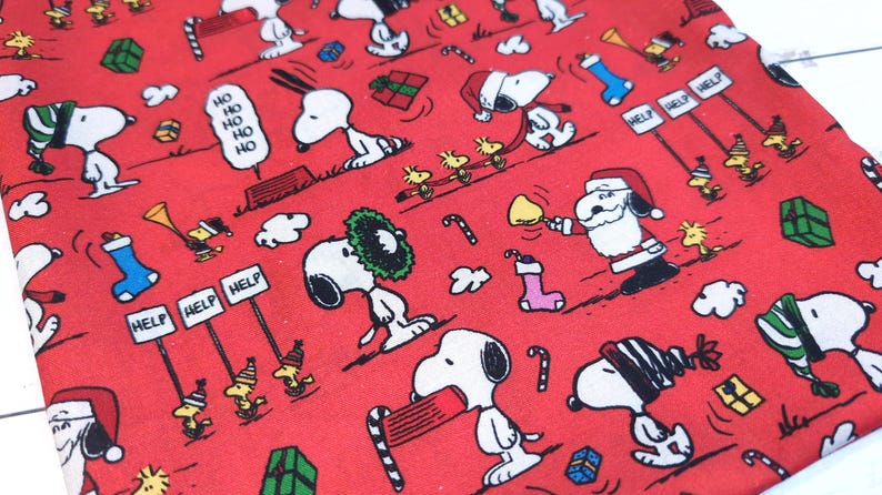 May include: Red fabric with a repeating pattern of Snoopy and Woodstock characters in Christmas scenes. Snoopy is wearing a Santa hat and scarf. The fabric also features candy canes, gifts, and the words "Ho Ho Ho" and "Help".