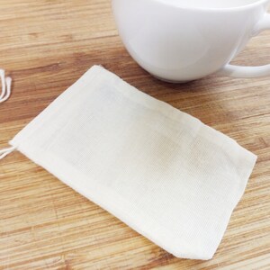 May include: A white cotton muslin tea bag with a drawstring closure. The tea bag is lying flat on a wooden surface.