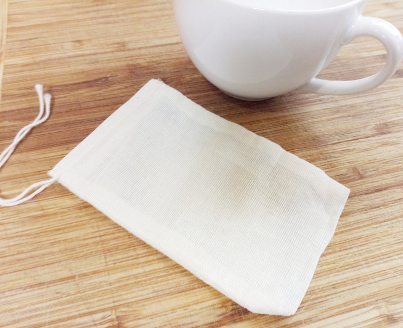 May include: A white cotton muslin tea bag with a drawstring closure. The tea bag is lying flat on a wooden surface.