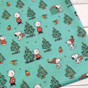 May include: Teal fabric featuring a repeating pattern of Snoopy, Charlie Brown, Christmas trees, and gift boxes. The characters are in their classic outfits, with the trees decorated with colorful ornaments. The fabric is suitable for holiday-themed crafts.