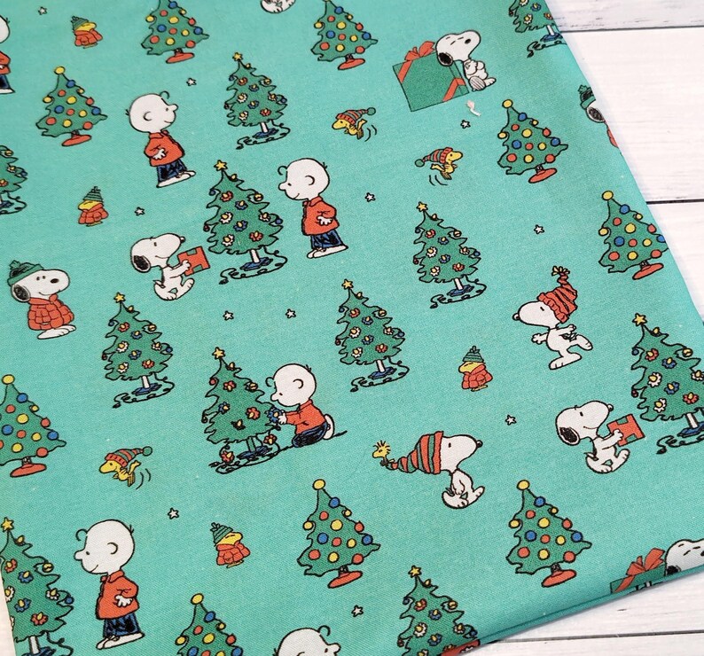 May include: Teal fabric featuring a repeating pattern of Snoopy, Charlie Brown, Christmas trees, and gift boxes. The characters are in their classic outfits, with the trees decorated with colorful ornaments. The fabric is suitable for holiday-themed crafts.