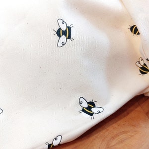 K&ouml;nnte beinhalten: Eine wei&szlig;e Stofftasche mit einem sich wiederholenden Muster aus schwarzen und gelben Bienen. Die Tasche ist gefaltet und liegt auf einer Holzoberfl&auml;che.