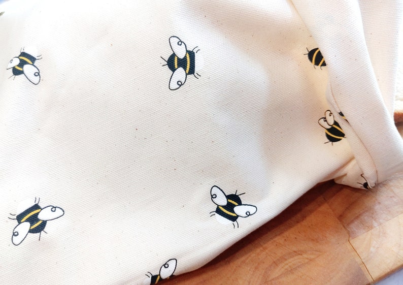 K&ouml;nnte beinhalten: Eine wei&szlig;e Stofftasche mit einem sich wiederholenden Muster aus schwarzen und gelben Bienen. Die Tasche ist gefaltet und liegt auf einer Holzoberfl&auml;che.