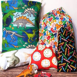 May include: Five colourful drawstring bags with various patterns. The bags are made of fabric and have a drawstring closure. The patterns include dinosaurs, numbers, clocks, and crayons. There is also a toy dinosaur and a toy car in the image.
