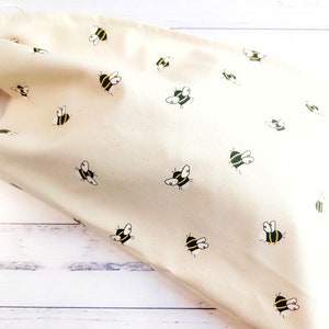 K&ouml;nnte beinhalten: Eine beige Kordeltasche mit einem Bienenmuster. Die Tasche ist aus Naturstoff gefertigt und hat ein schlichtes Design.