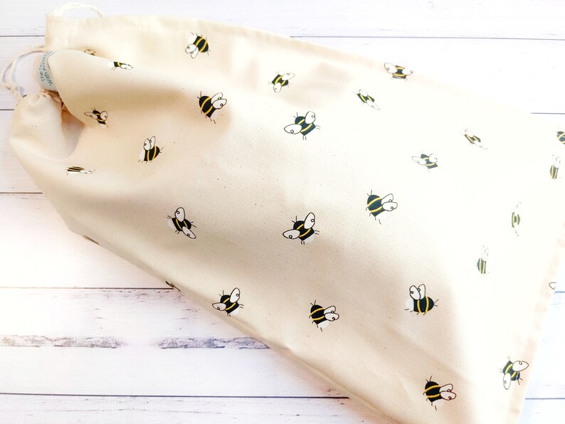 K&ouml;nnte beinhalten: Eine beige Kordeltasche mit einem Bienenmuster. Die Tasche ist aus Naturstoff gefertigt und hat ein schlichtes Design.