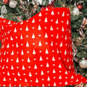 May include: A red cushion cover featuring a repeating pattern of white Christmas trees and snowflakes. The cover is made of a soft fabric and is shown in front of a decorated Christmas tree.