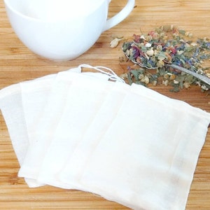 May include: Four white muslin tea bags, stacked on a wooden surface, with a spoon of dried herbs and flowers scattered nearby.