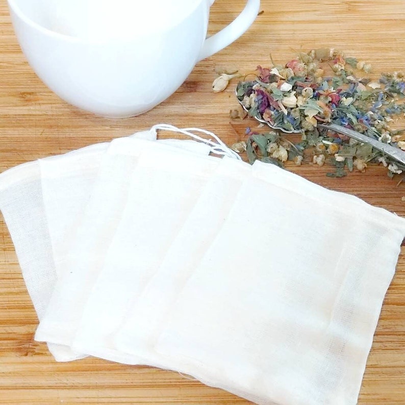 May include: Four white muslin tea bags, stacked on a wooden surface, with a spoon of dried herbs and flowers scattered nearby.