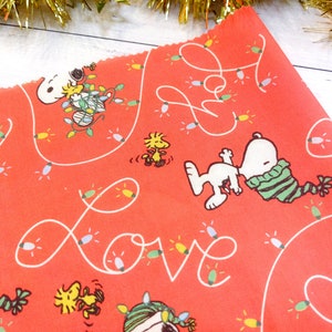 May include: Red fabric featuring Snoopy and Woodstock characters with Christmas lights and the word "Love" in white. Snoopy is wearing a green and white striped scarf. The fabric is likely for crafting or holiday decor.