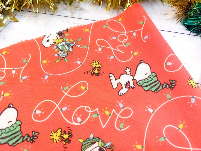 May include: Red fabric featuring Snoopy and Woodstock characters with Christmas lights and the word "Love" in white. Snoopy is wearing a green and white striped scarf. The fabric is likely for crafting or holiday decor.